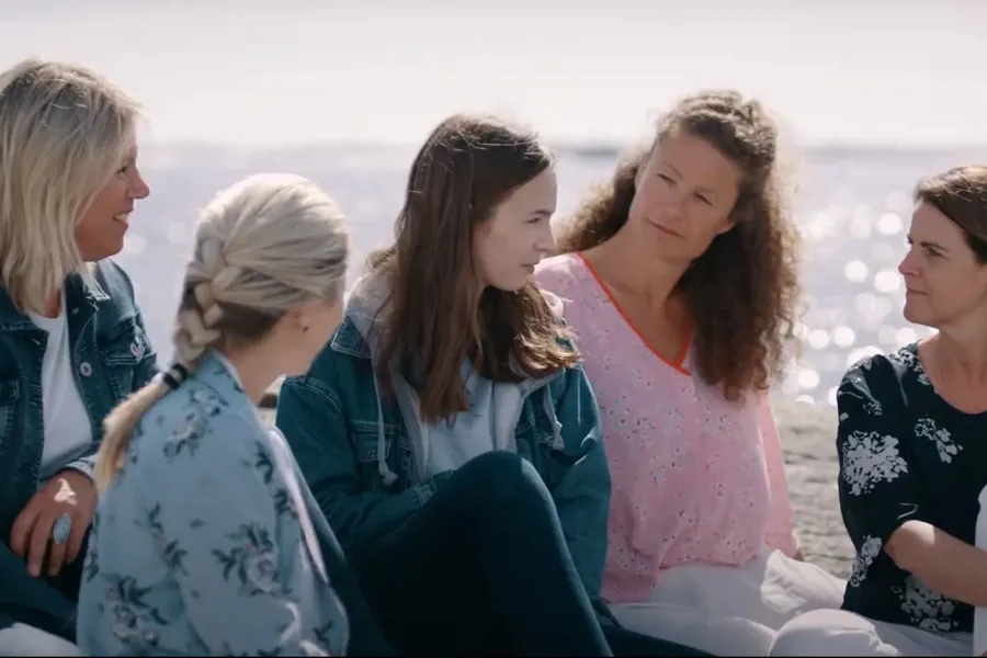 En ungdom sitter ved kysten og snakker til fire voksne rundt. Bilde.