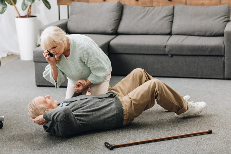 En eldre mann har falt og ligger på gulvet. En eldre dame holder en telefon og hjelper. Bilde.