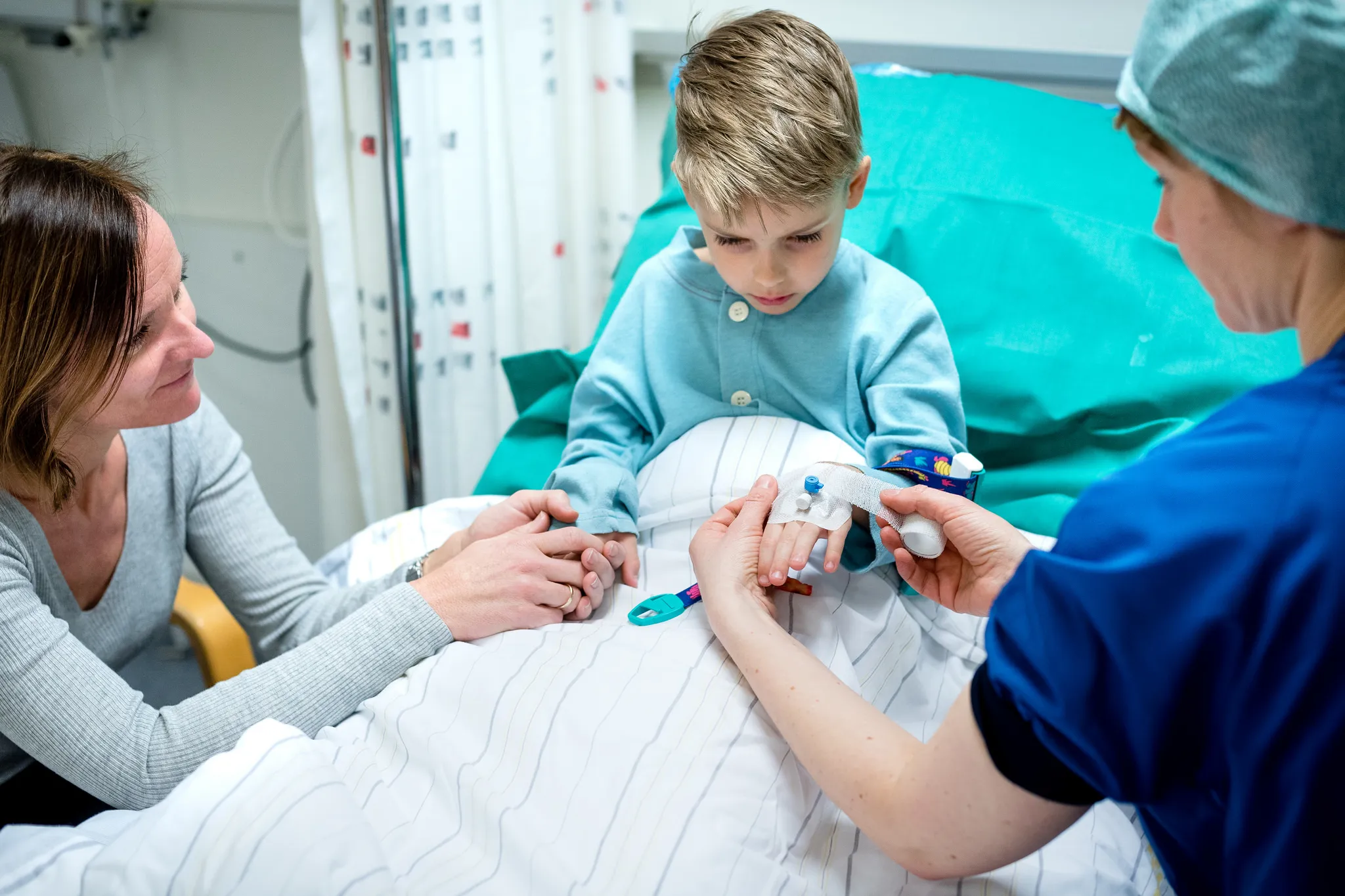 Mor og sønn ser når en sykepleier i blå klær setter inn en veneflon i guttens hånd. Foto.