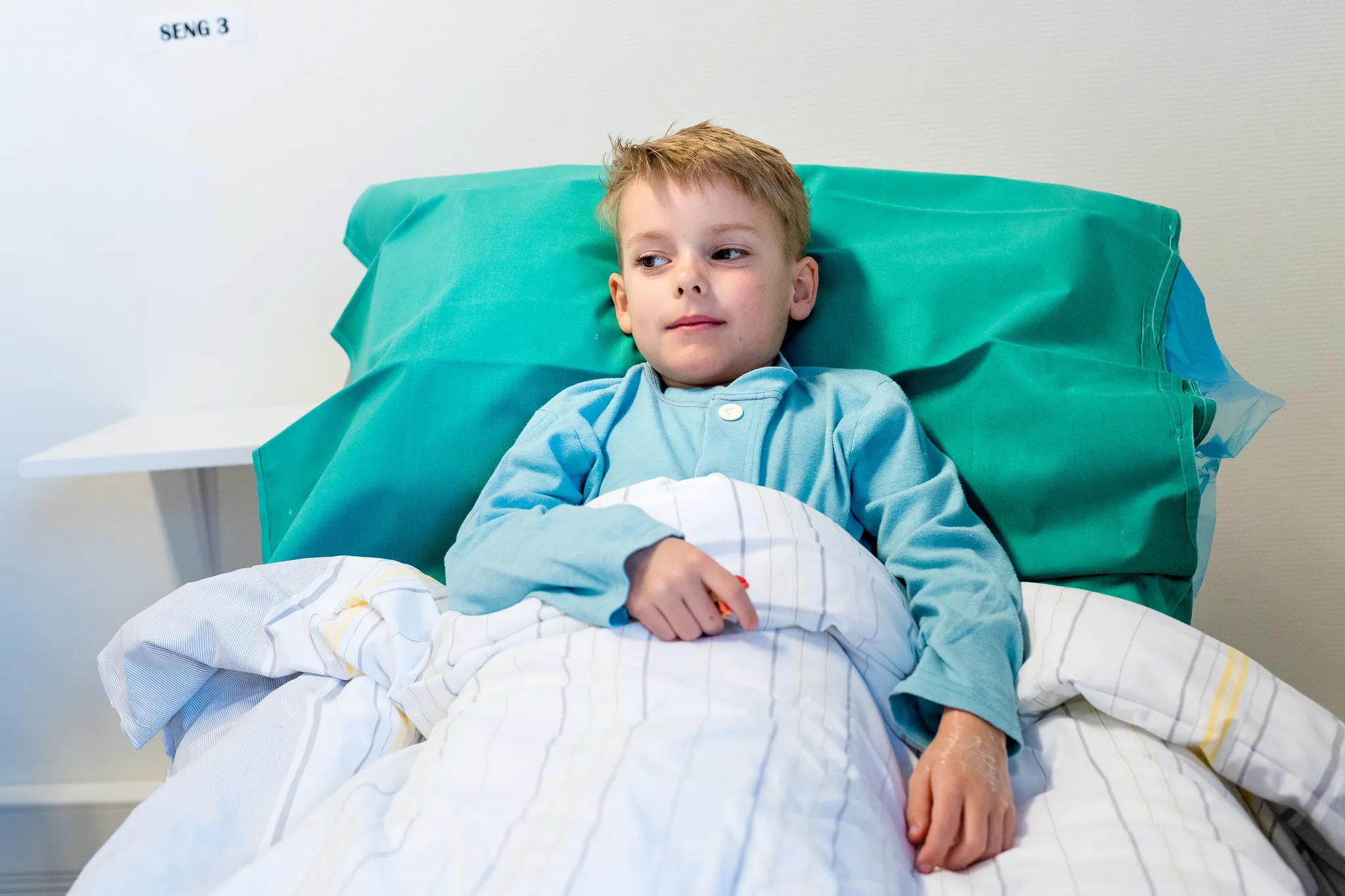 Liten gutt med sykehusklær ligger i en sykehusseng. Smiler forsiktig. Foto.