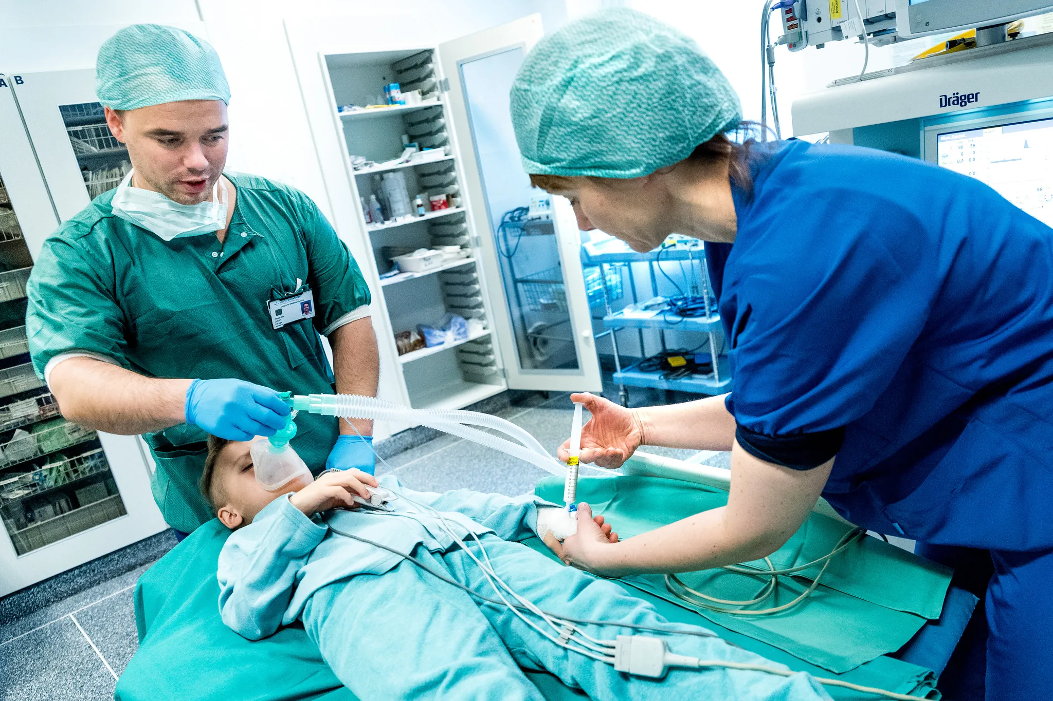 Liten gutt i sykehusklær ligger på en operasjonsseng. Anestesilege og anestesisykepleier gir han sovemedisin. Foto.