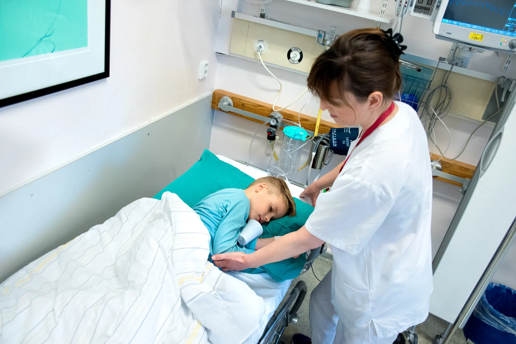 Liten gutt i sykehusklær ligger og sover i en sykehusseng. Sykepleier i hvite klær passer på.
