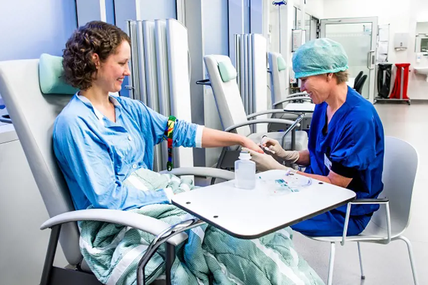 Sykepleier i blå klær legger veneflon på dagkirurgisk pasient som sitter i en stol. Foto.