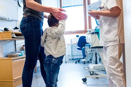 Barn som står på en vekt imens mor holder i hendene. Sykepleier smiler til barnet. Foto.